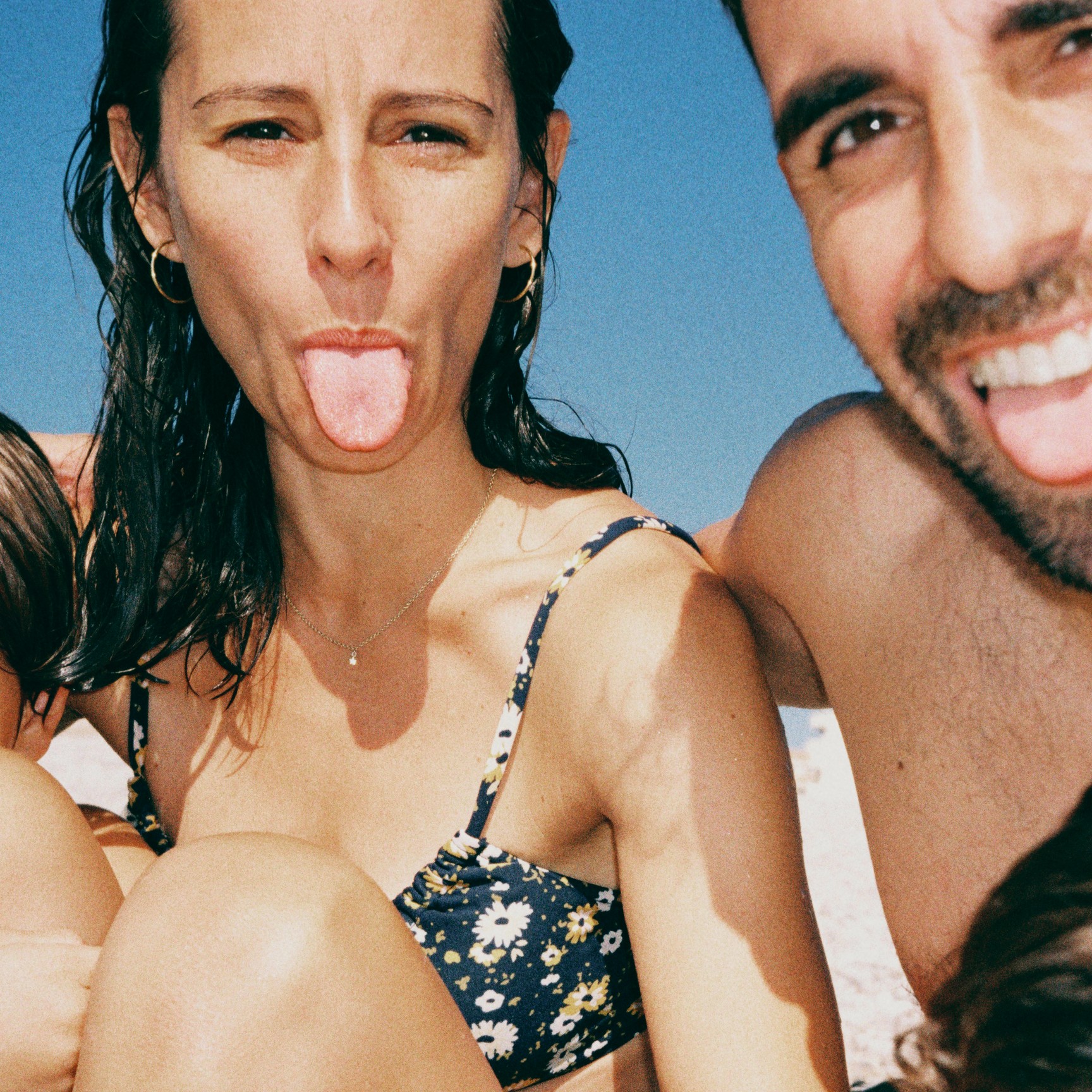 A close-up, family selfie at the beach. A man and woman smile and stick their tongues out at the camera, with their children partially in the shot.