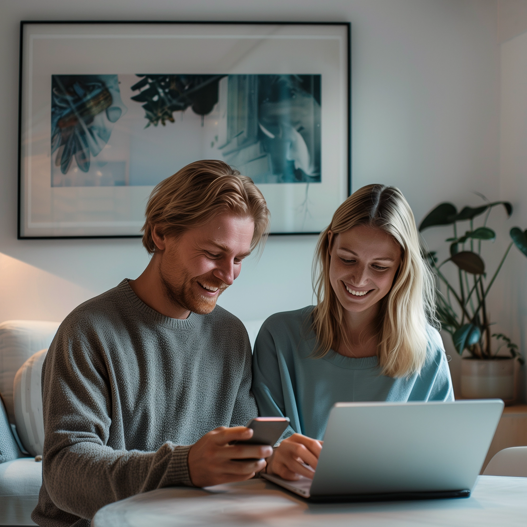 A smiling man and woman sit together at a table in their home, looking at a smartphone and a laptop.