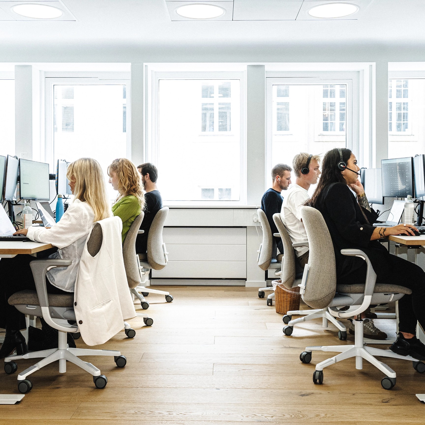 En vy längs gången i ett ljust, öppet kontorslandskap, där anställda arbetar vid sina datorer i två rader. Flera av medarbetarna bär headset.