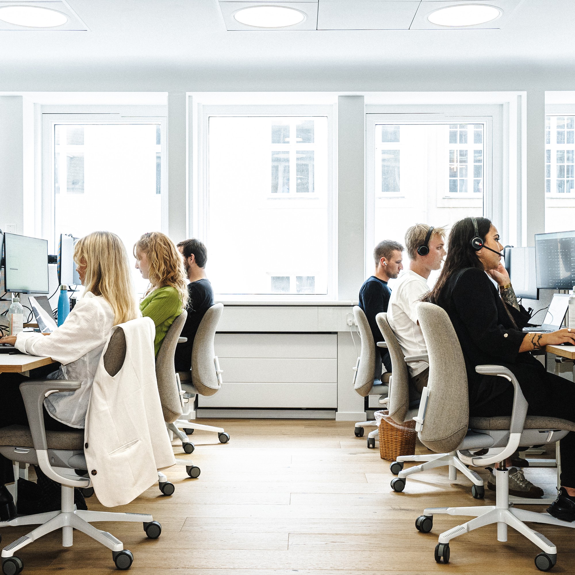 A view down the aisle of a bright, open-plan office, where employees are working at their computers in two rows. Several staff members are wearing headsets.