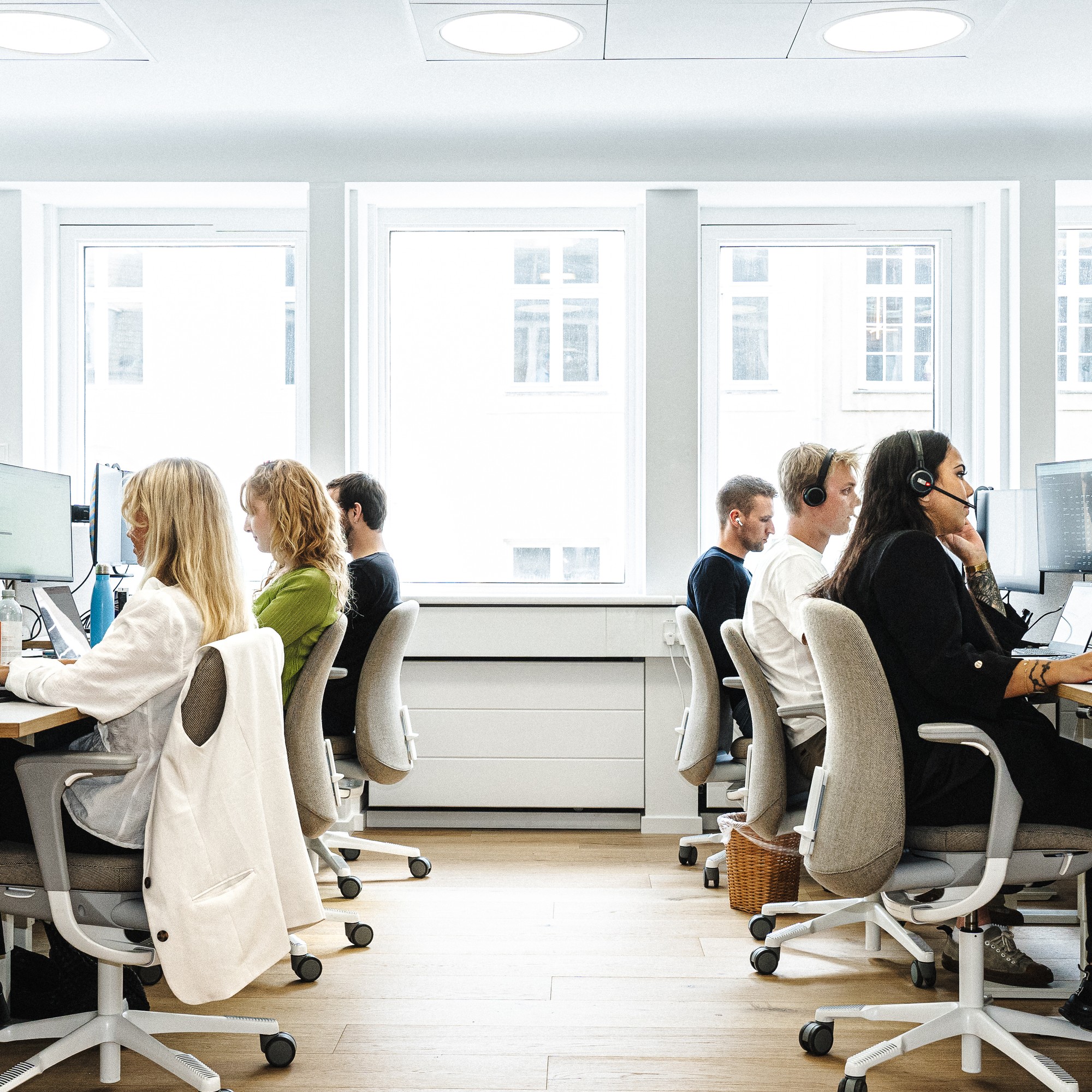 Utsikt nedover midtgangen i et lyst, åpent kontorlandskap, der ansatte jobber på datamaskinene sine i to rader. Flere av de ansatte har på seg headset.