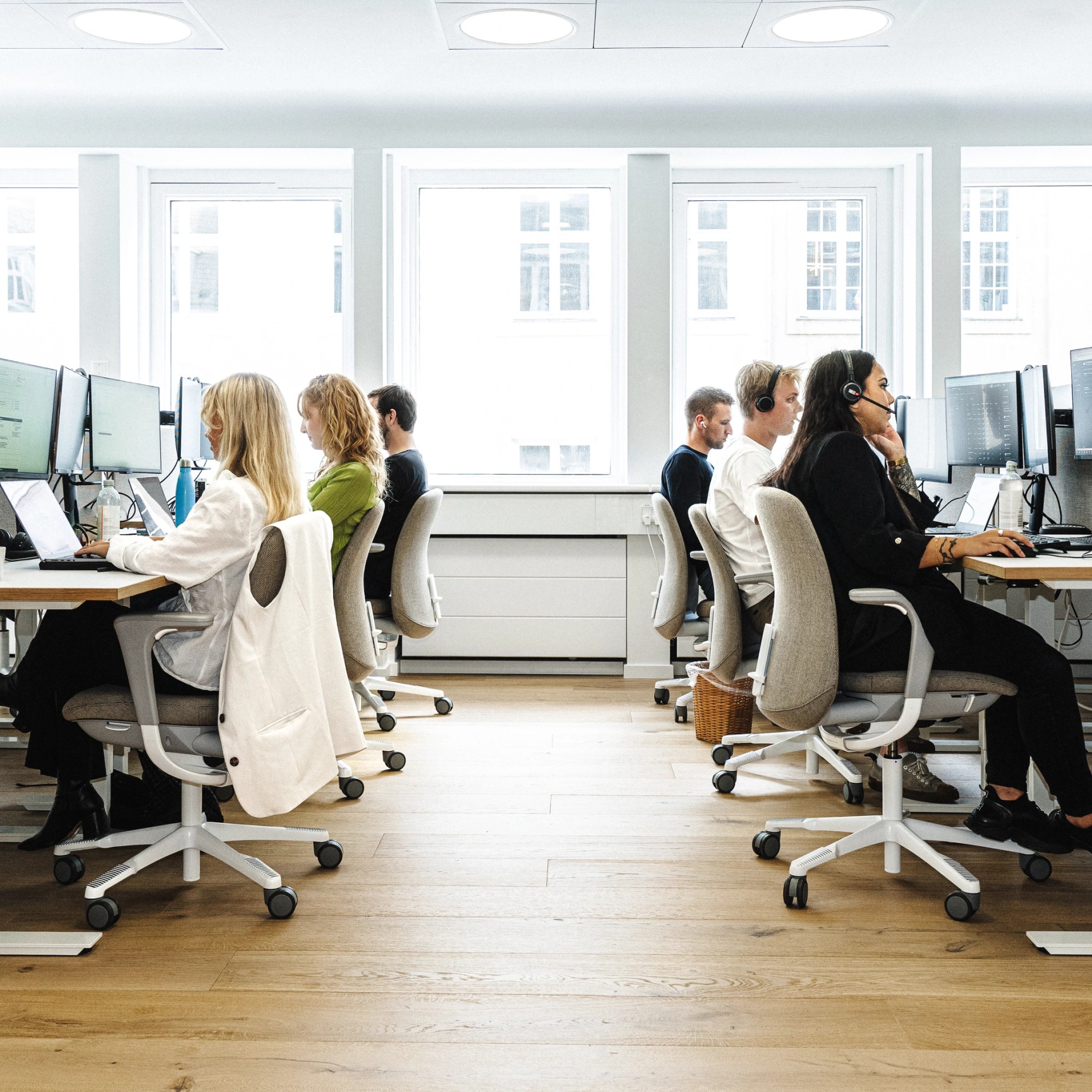 Udsigt ned ad gangen i et lyst, åbent kontorlandskab, hvor medarbejdere arbejder ved deres computere i to rækker. Flere af medarbejderne bærer headsets.