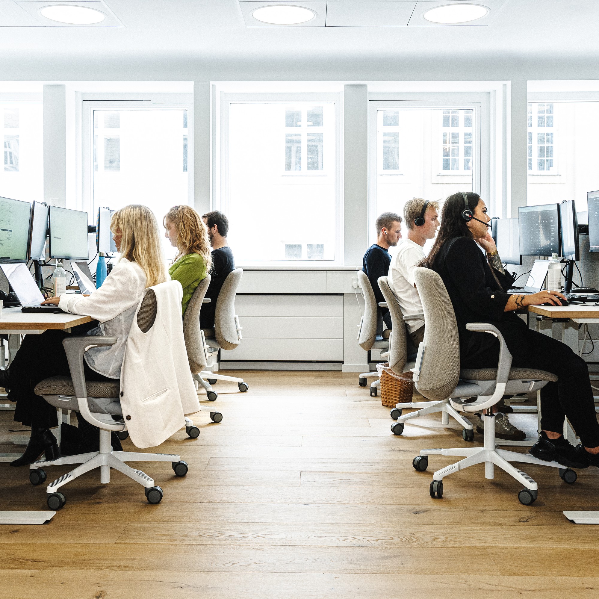 Udsigt ned ad gangen i et lyst, åbent kontorlandskab, hvor medarbejdere arbejder ved deres computere i to rækker. Flere af medarbejderne bærer headsets.