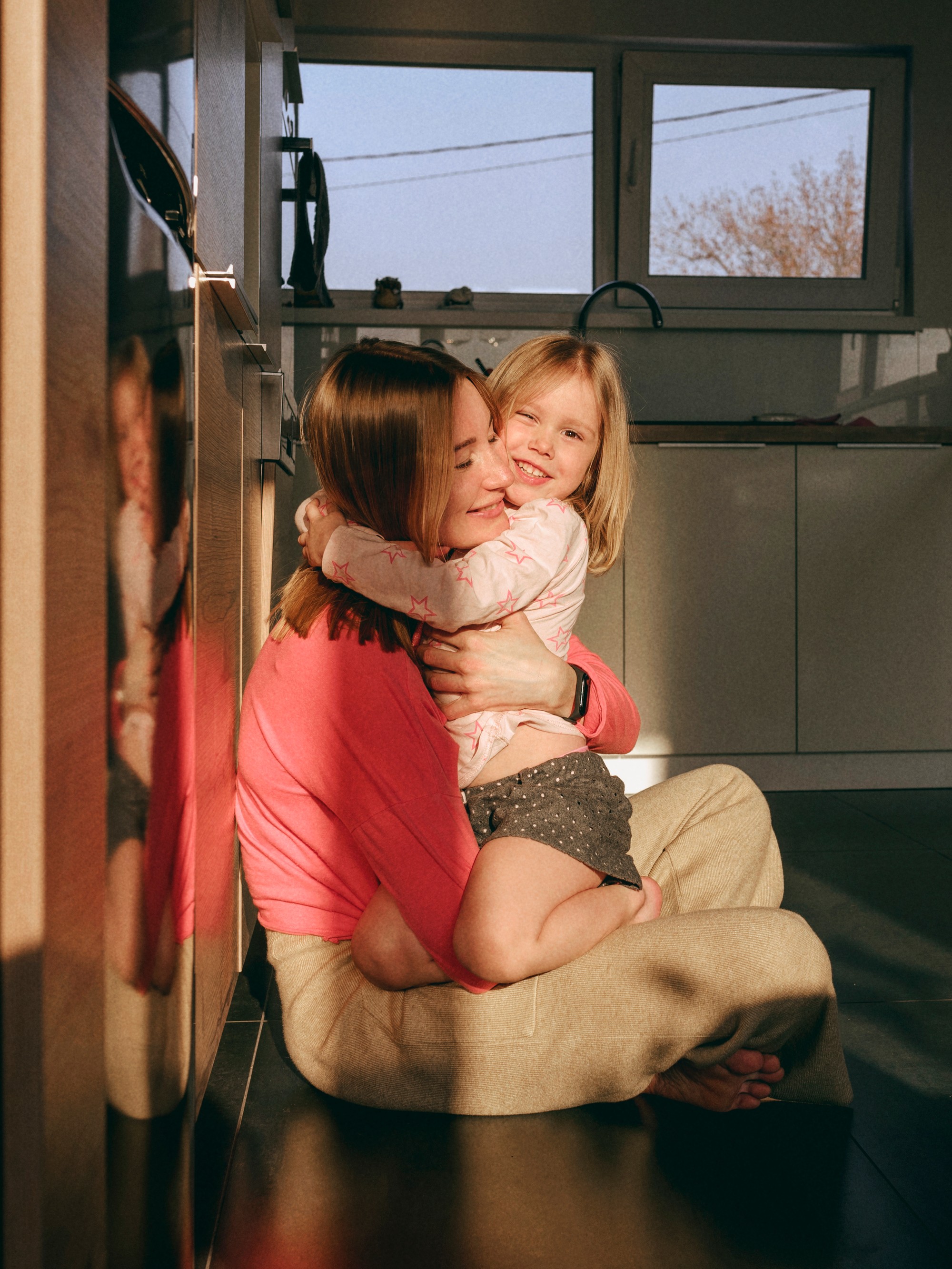 En mor og hendes lille datter deler et gladt kram, mens de sidder på gulvet i et solbeskinnet køkken.