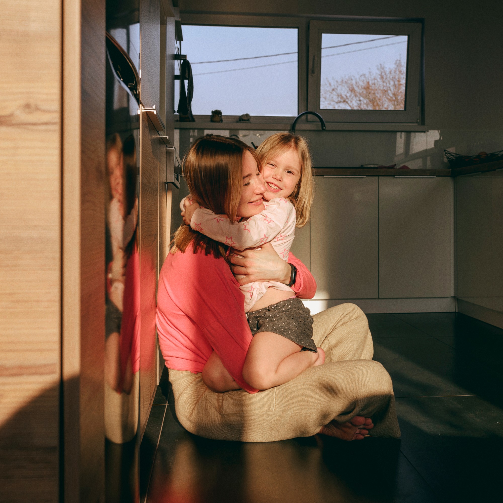 En mamma och hennes lilla dotter delar en glad kram medan de sitter på golvet i ett solbelyst kök.
