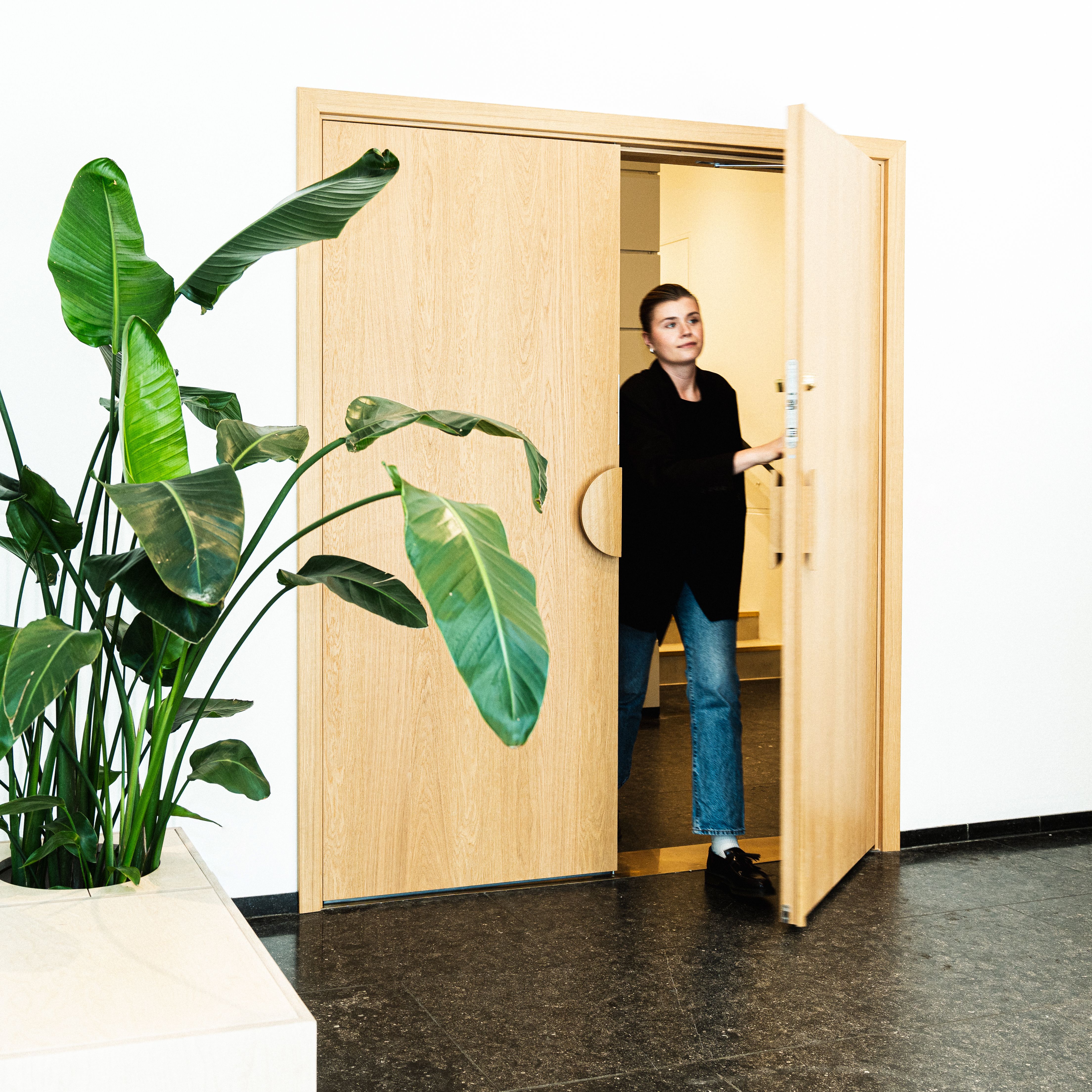 En person går gjennom et sett med lyse tredører i en moderne, minimalistisk kontor-lobby. En stor, grønn bladplante i en hvit plantekasse står ved siden av døråpningen.