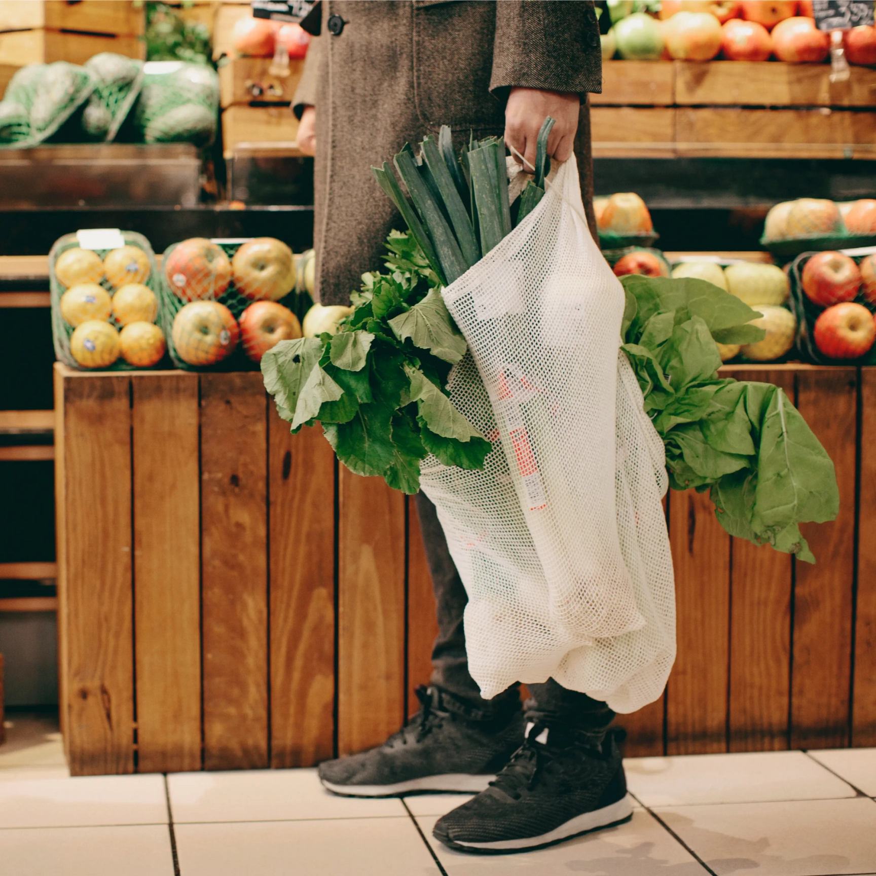 En person står i en mataffär och håller i en återanvändbar nätkasse fylld med färska grönsaker som purjolök och bladgrönsaker.