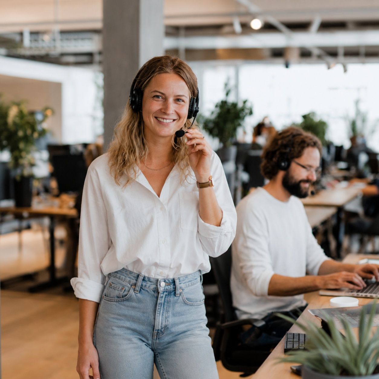 En venlig kvindelig kundeservicemedarbejder med et headset smiler, mens hun arbejder ved sin computer på et lyst, moderne kontor.