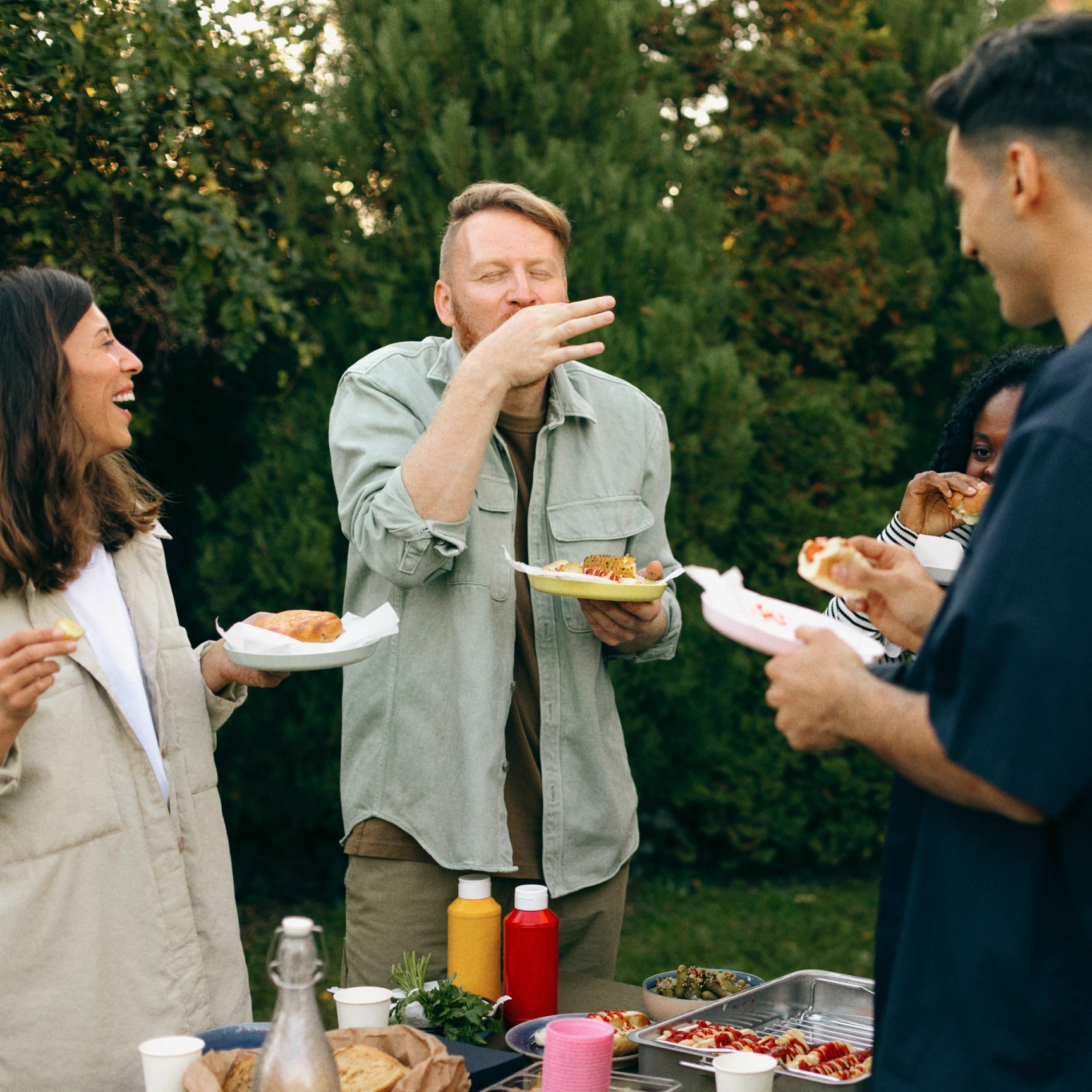 En grupp med olika vänner skrattar och äter tillsammans vid en utomhusgrillfest i en trädgård.