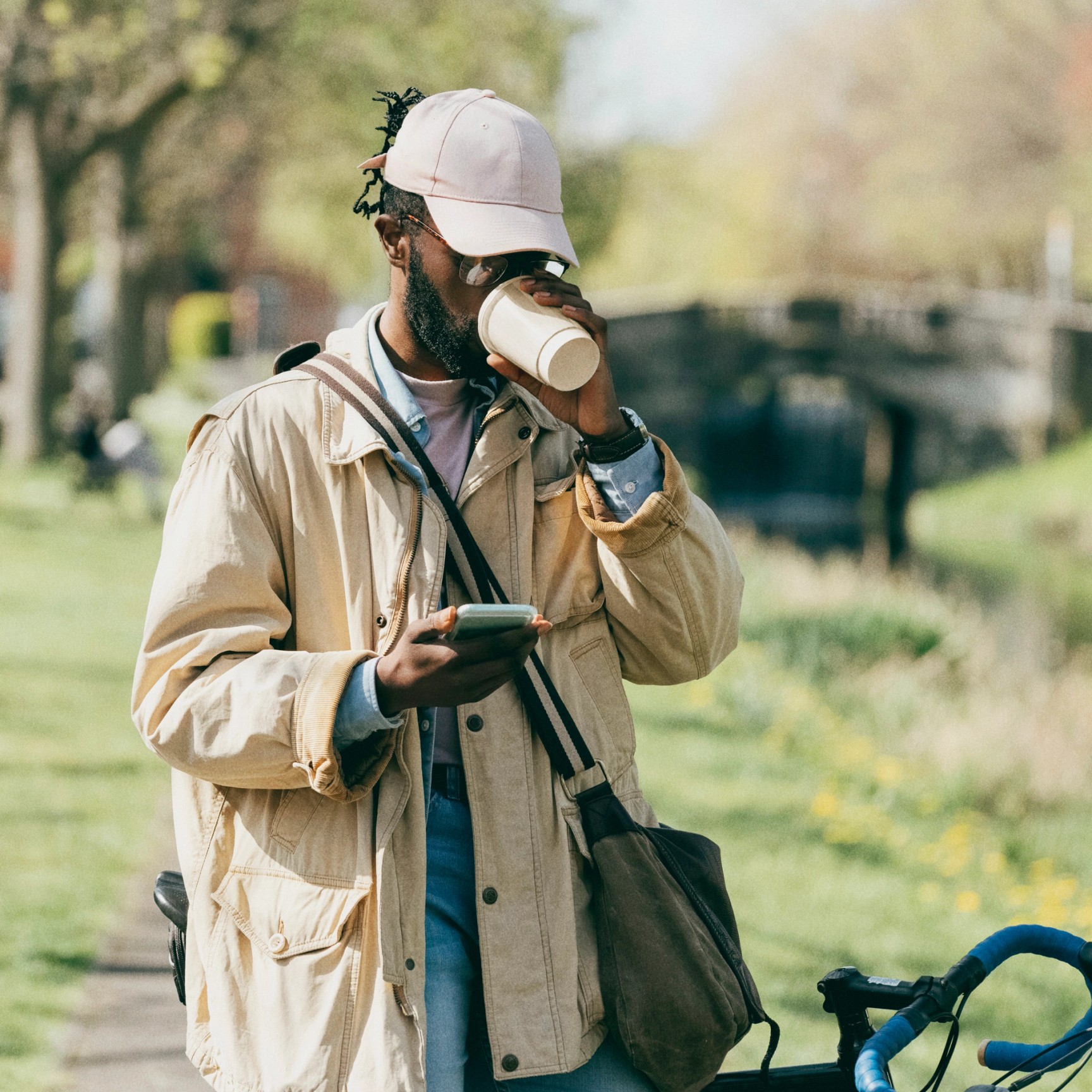A man with a bicycle stands in a park, looking at his smartphone while drinking coffee.