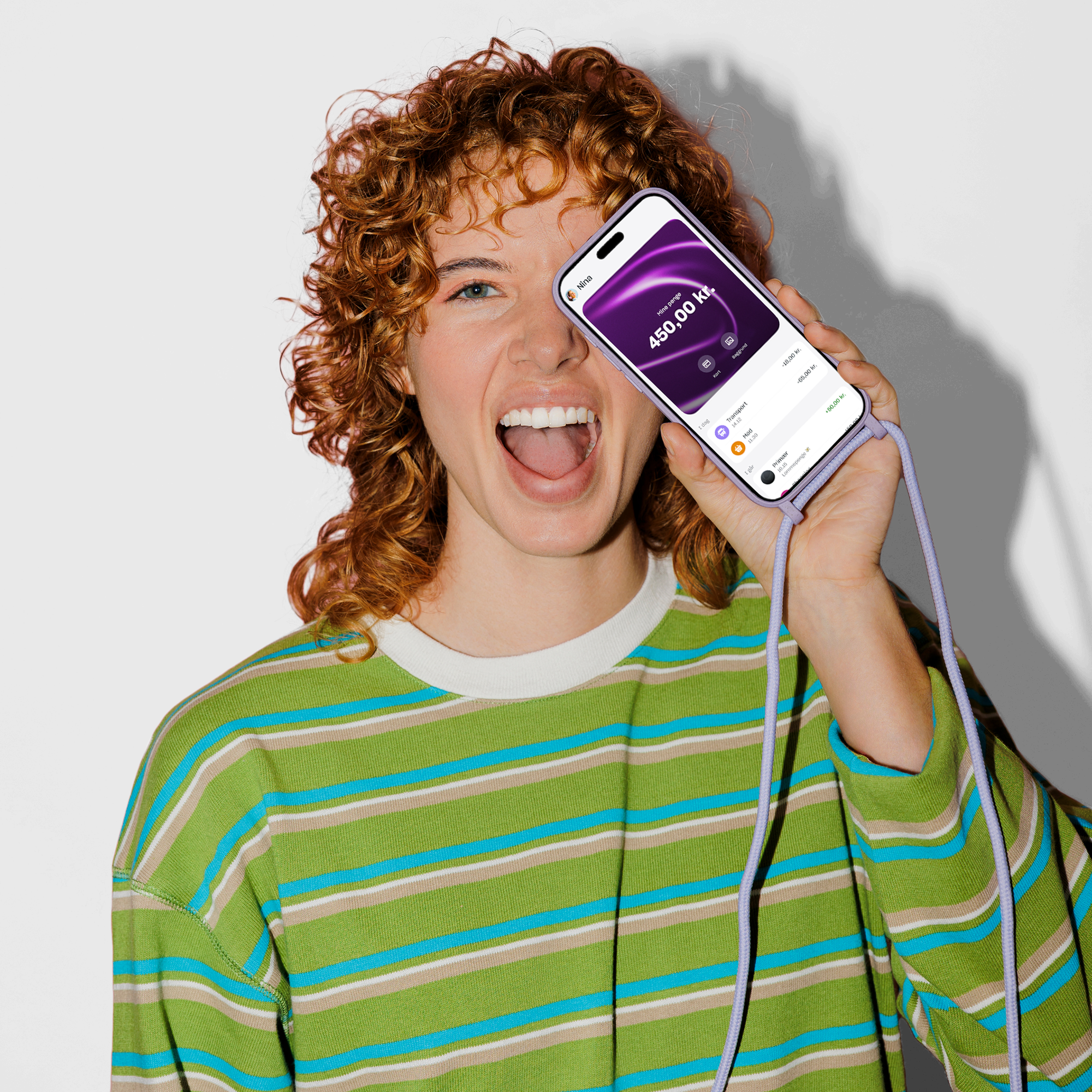 En glad ung kvinde med krøllet rødt hår holder en smartphone med en lilla snor op til ansigtet, som viser en saldo på 450,00 kr. på skærmen.