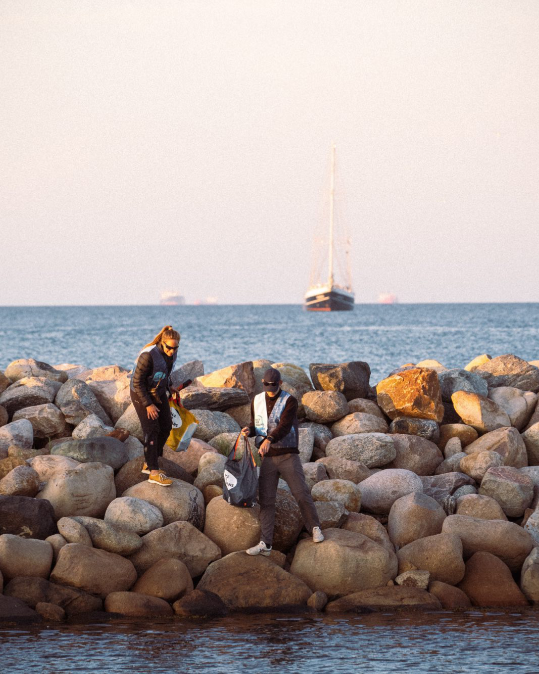 Två volontärer samlar skräp i en påse medan de står på en stenig pir, som en del av ett kuststädnings-evenemang.