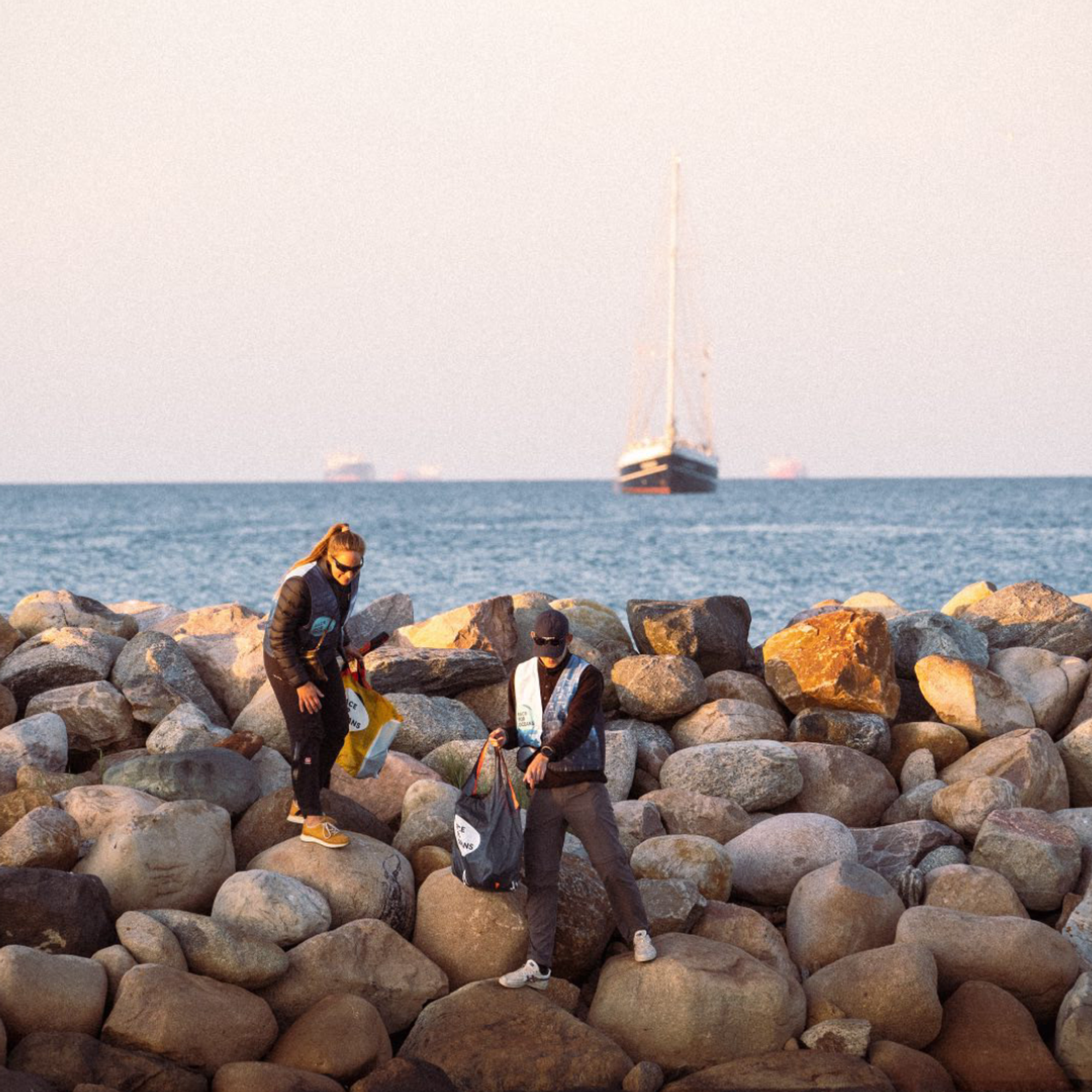 Två volontärer samlar skräp i en påse medan de står på en stenig pir, som en del av ett kuststädnings-evenemang.