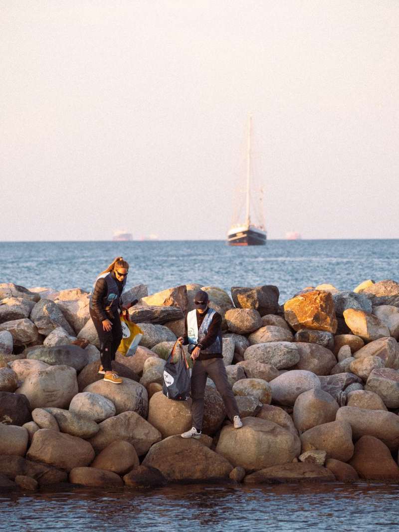 Two volunteers collect trash in a bag while standing on a rocky sea wall, as part of a coastal cleanup event.