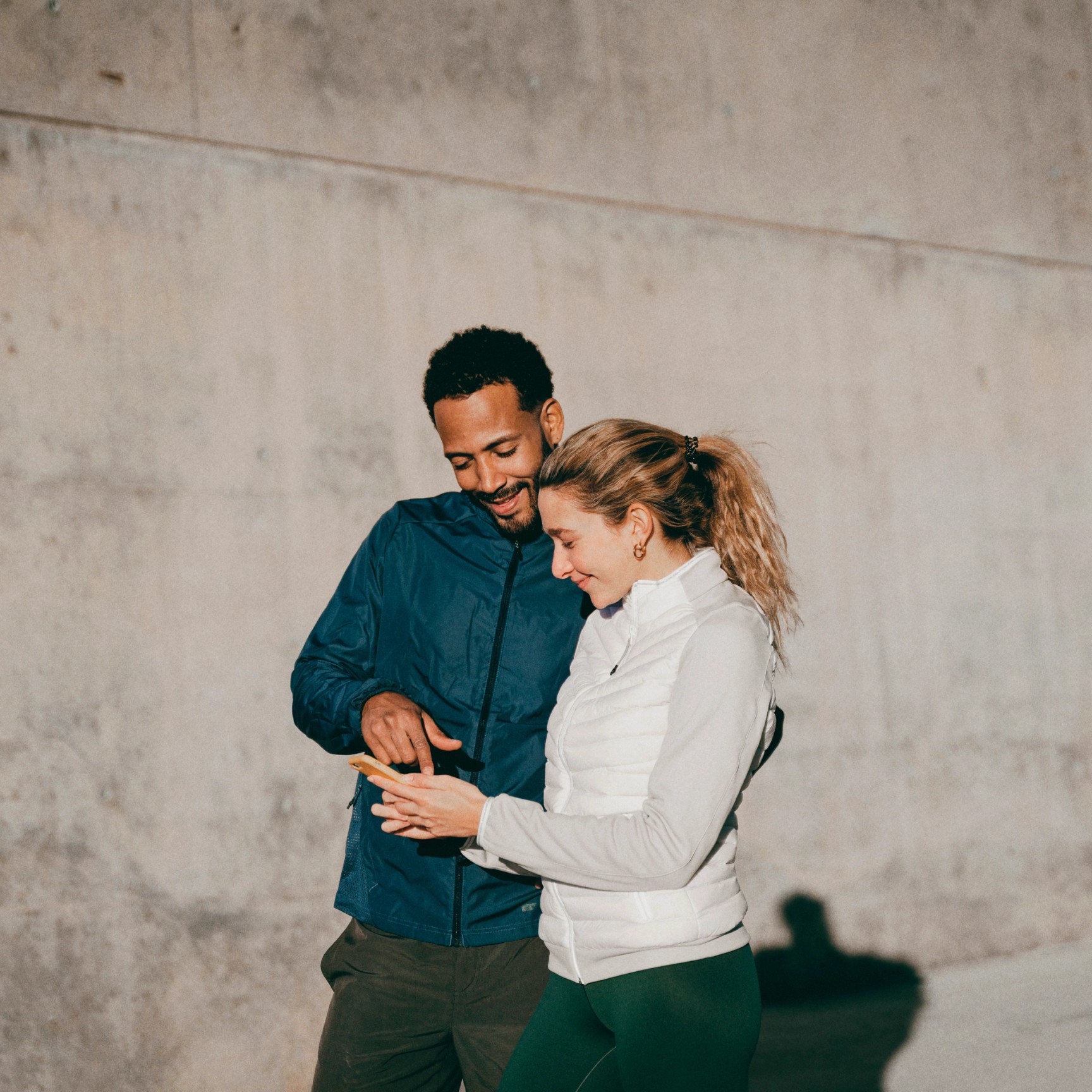 En mann og en kvinne i uformelle treningsjakker står foran en betongvegg og smiler mens de ser på en smarttelefon sammen. Mannen peker på telefonskjermen, som kvinnen holder.
