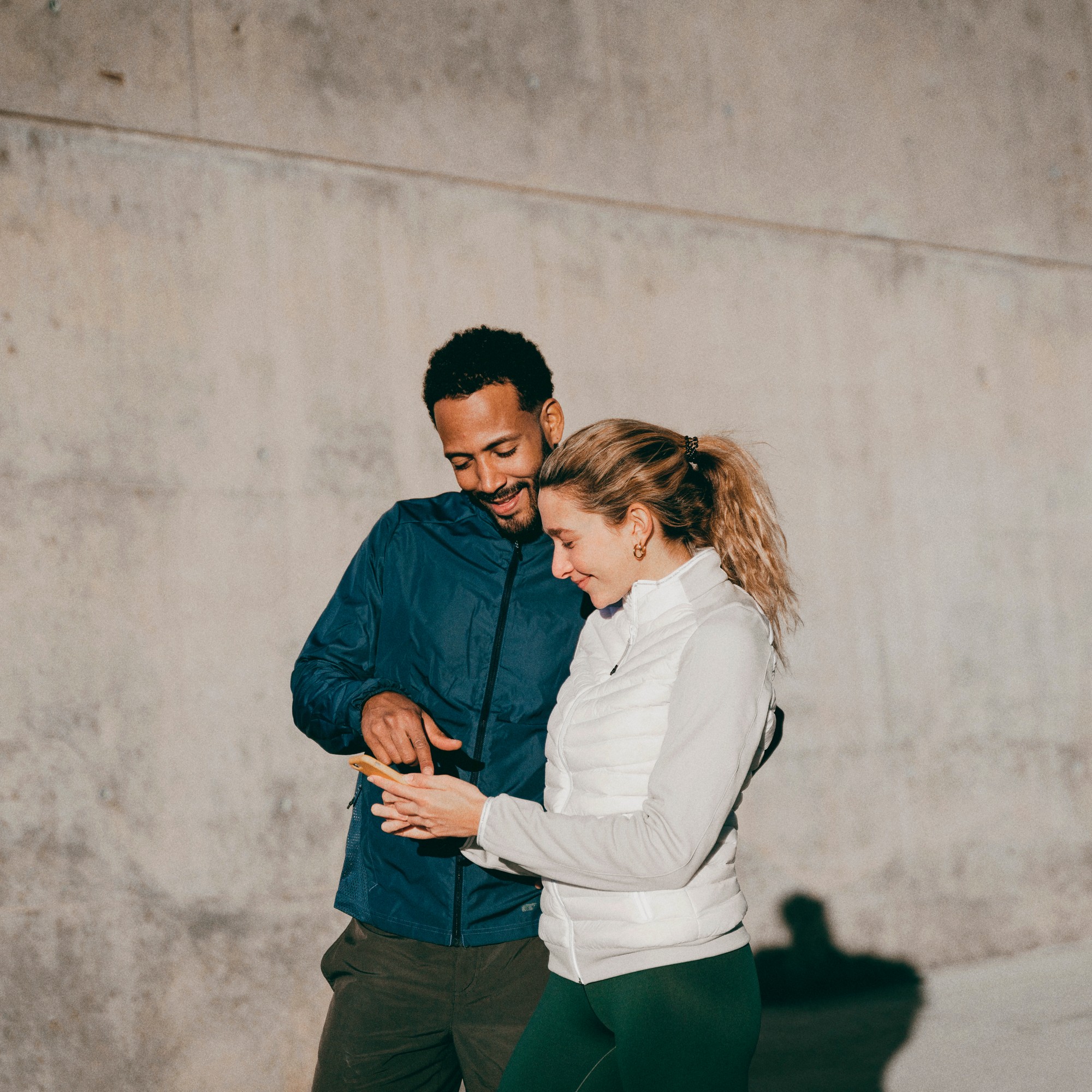 A man and a woman in casual athletic jackets look at a smartphone together. The man is pointing at the screen as they stand in front of a concrete wall.