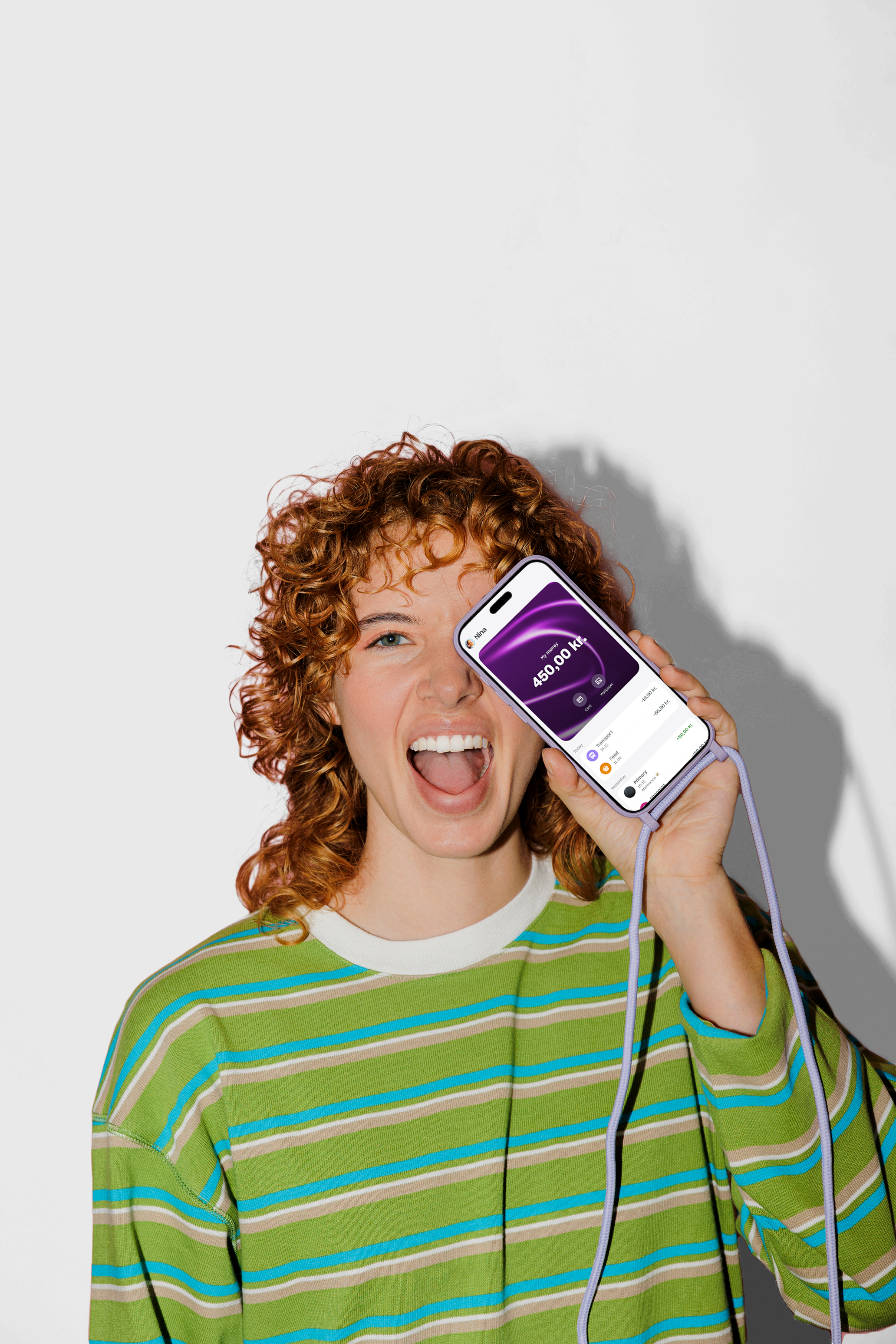 A joyful young woman with curly red hair holds a smartphone with a purple lanyard up to her face, showing a balance of 450.00 kr. on the screen.