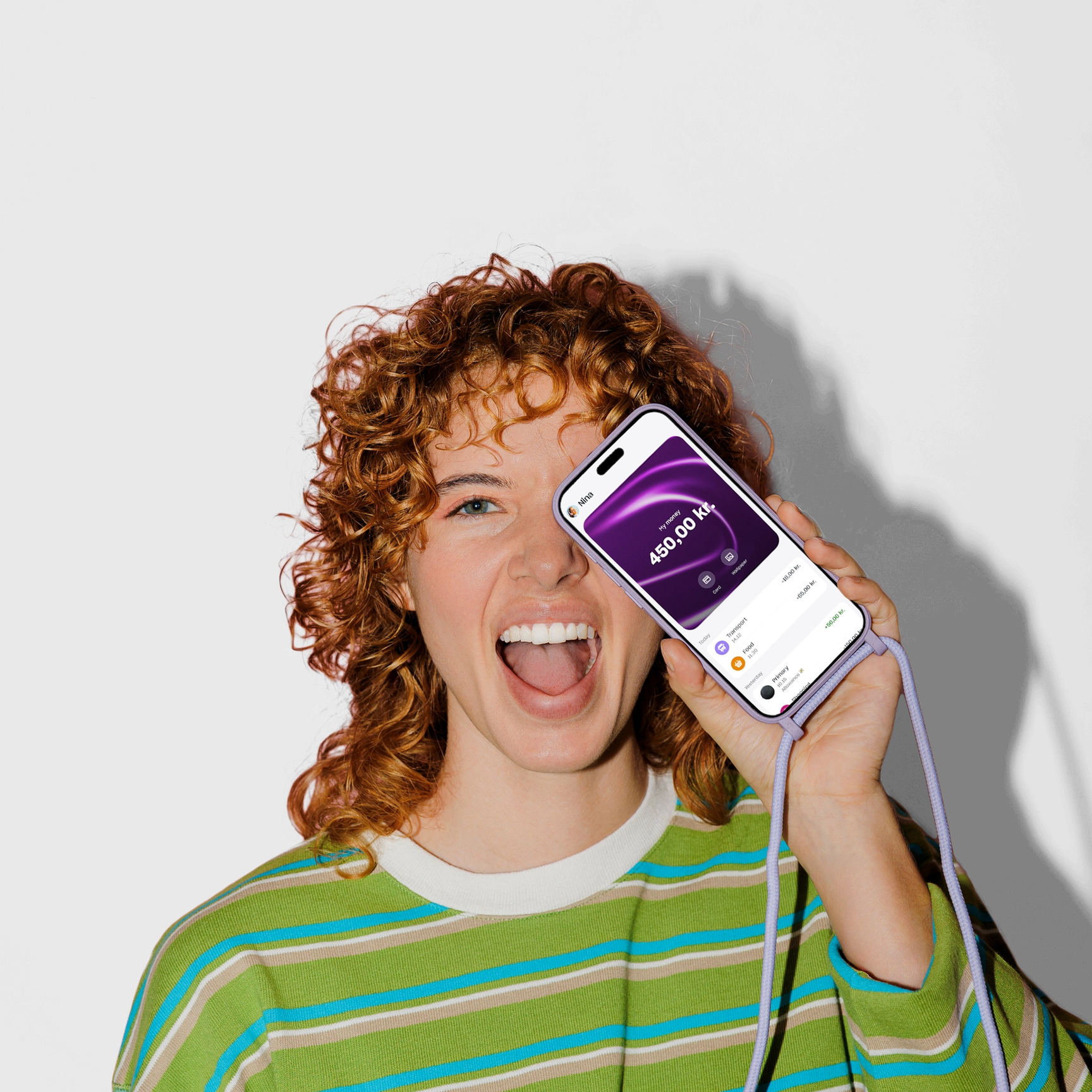 A joyful young woman with curly red hair holds a smartphone with a purple lanyard up to her face, showing a balance of 450.00 kr. on the screen.