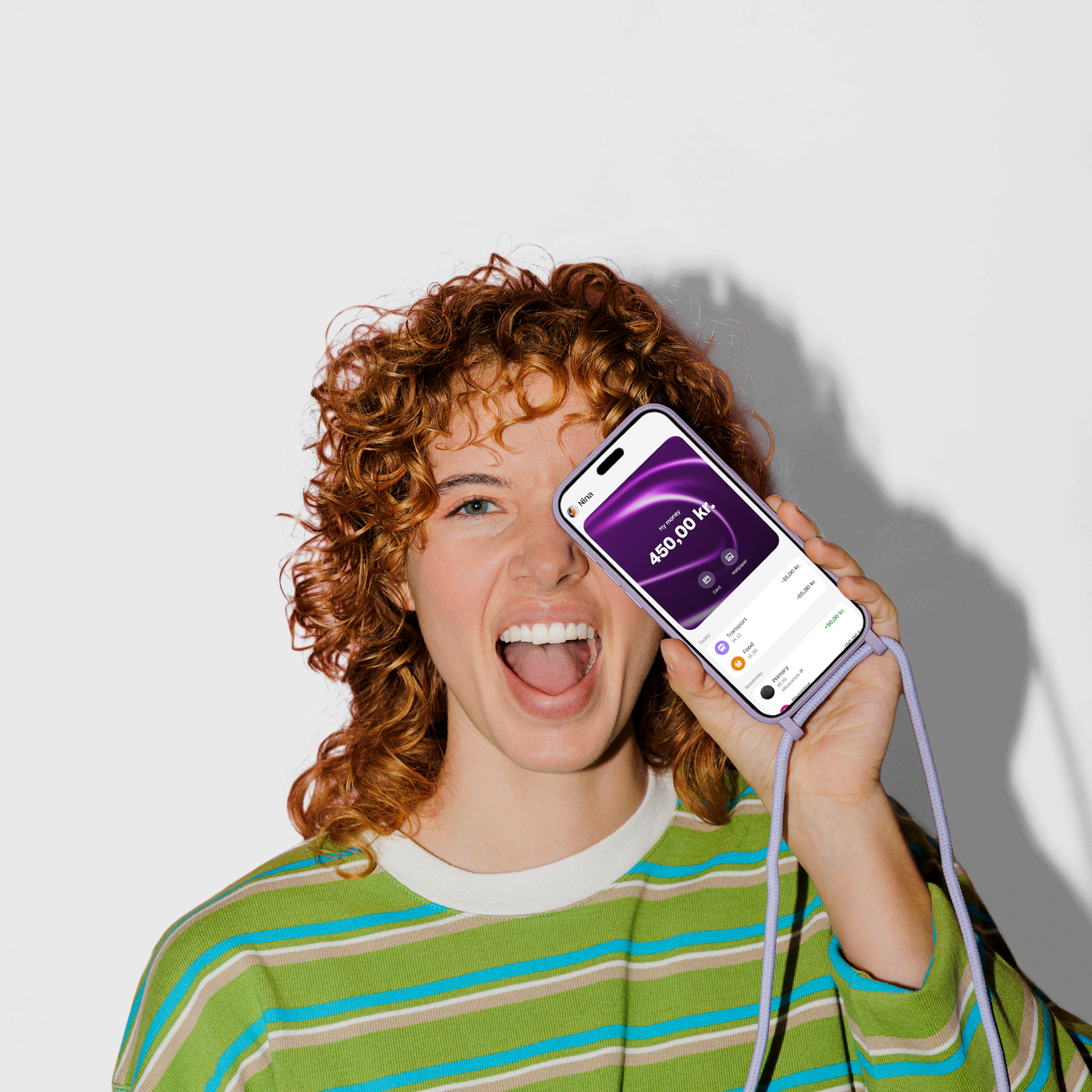 A joyful young woman with curly red hair holds a smartphone with a purple lanyard up to her face, showing a balance of 450.00 kr. on the screen.