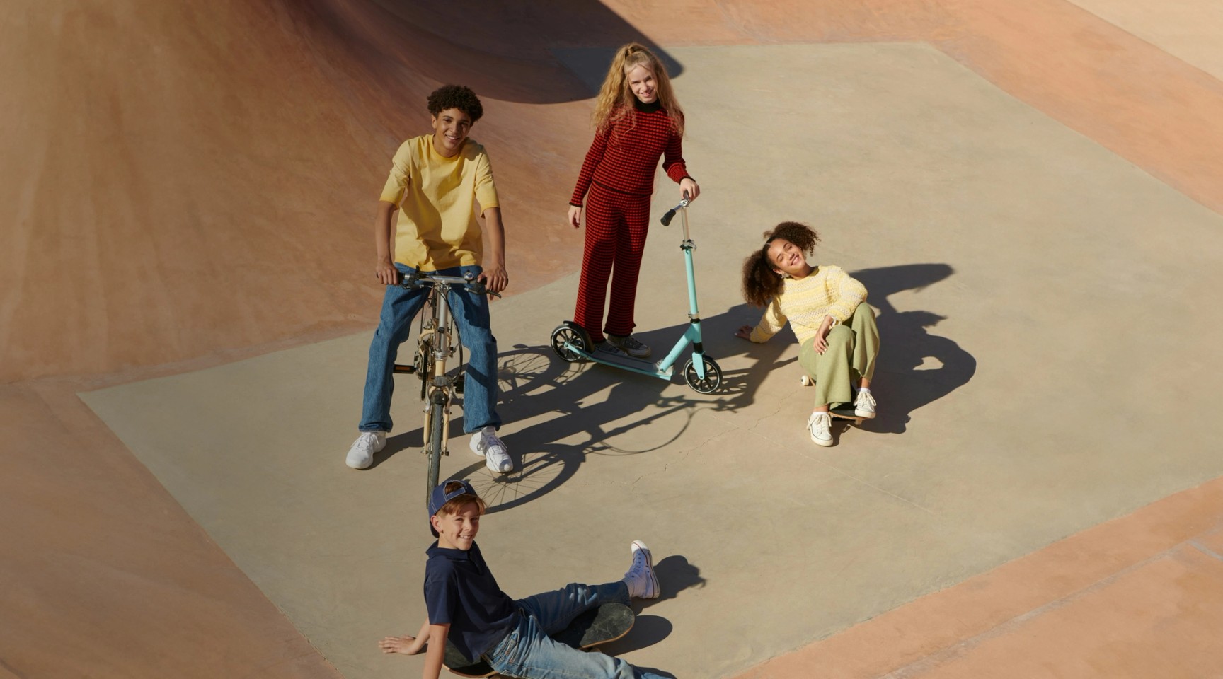 En mångfaldig grupp på fyra barn och tonåringar hänger i en skatepark i betong med en cykel, en sparkcykel och en skateboard.