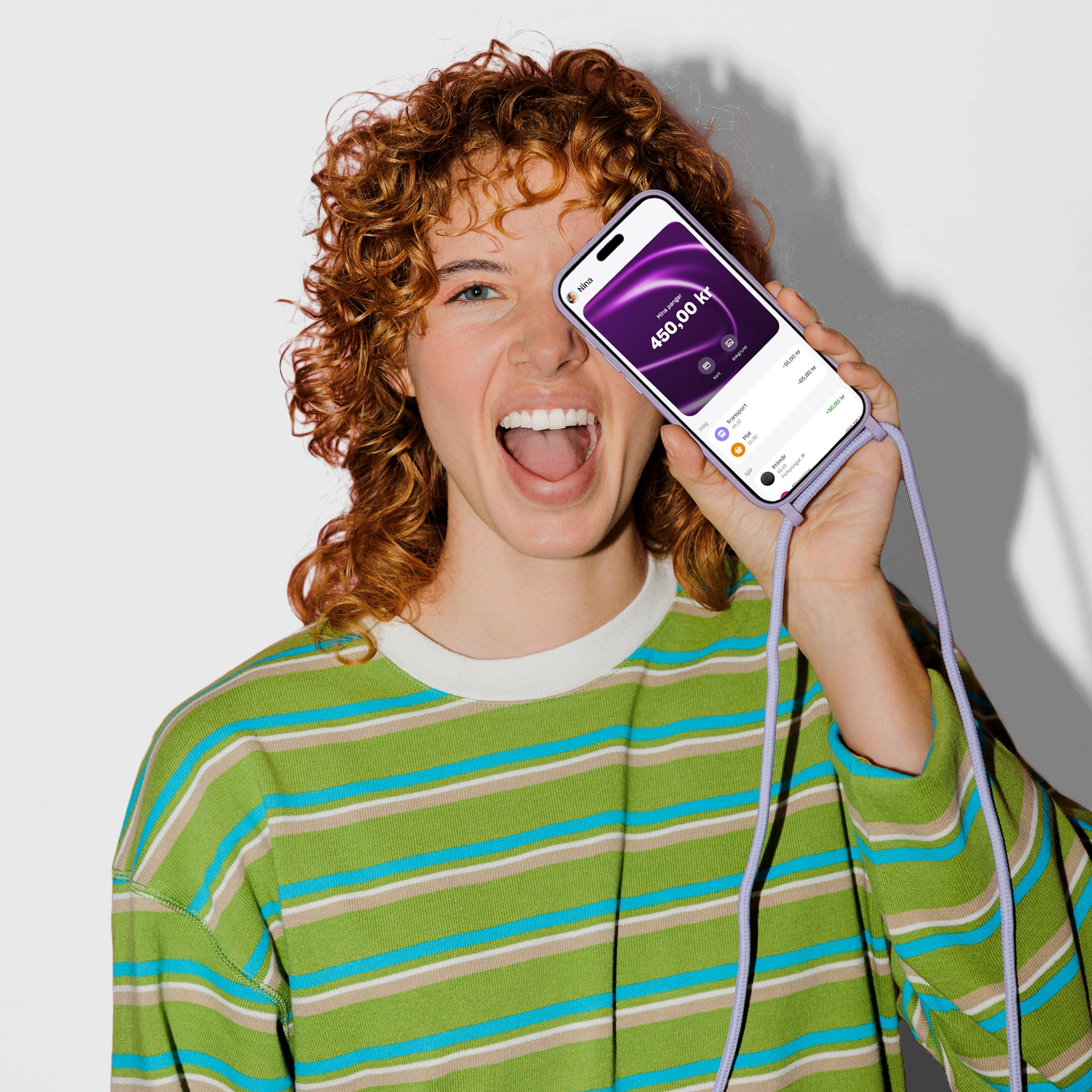 En glad ung kvinna med lockigt rött hår håller upp en smartphone med ett lila snöre framför ansiktet, som visar ett saldo på 450,00 kr på skärmen.