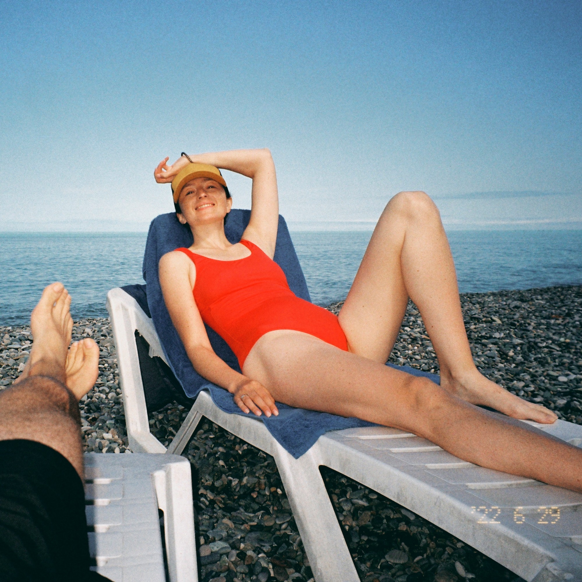 A woman in a red swimsuit and yellow cap smiles while relaxing on a lounger on a pebble beach, with the sea behind her.