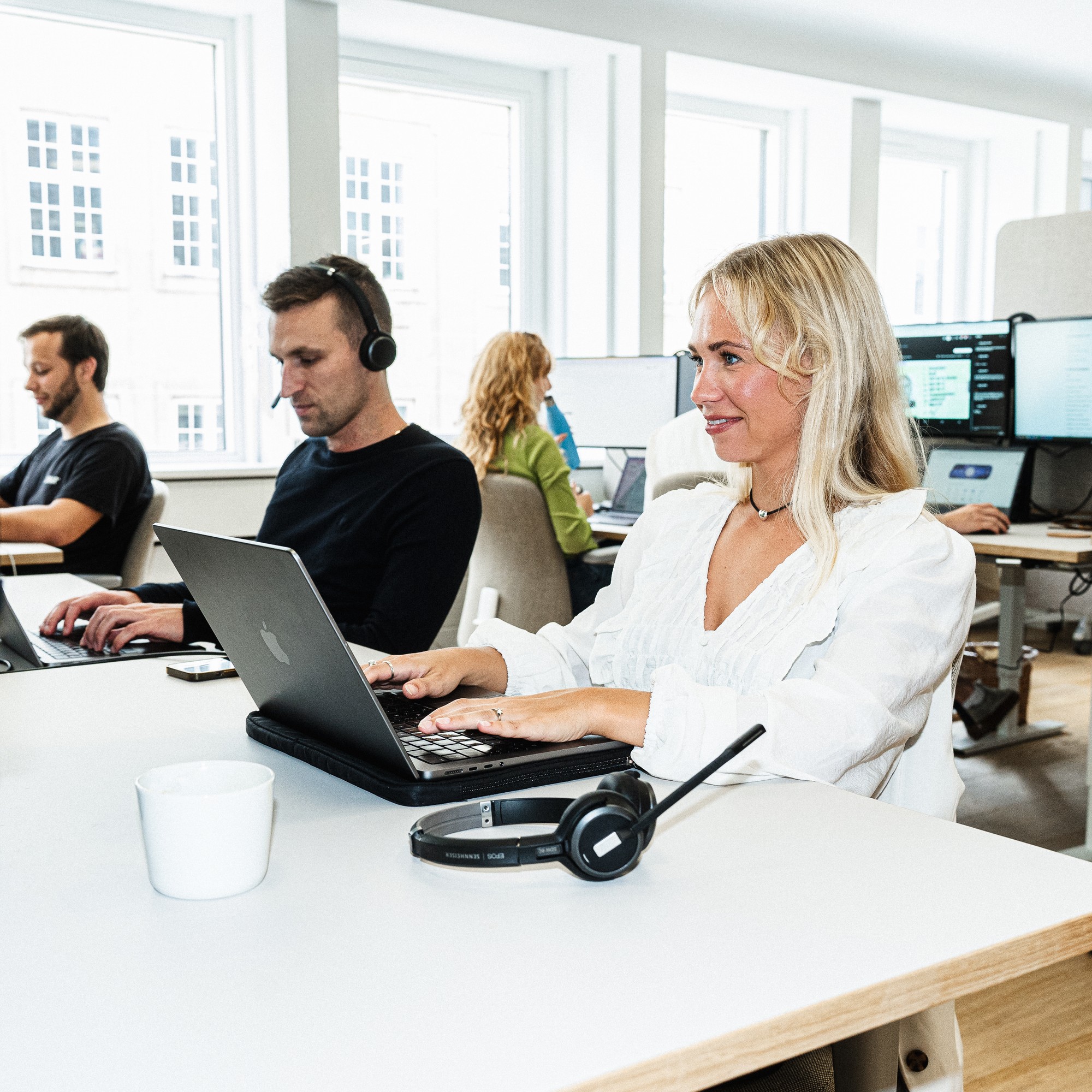 Et team af folk, der arbejder på bærbare computere på et lyst, moderne kontor. En blond kvinde i forgrunden smiler, mens hun taster, med et headset liggende på sit skrivebord.