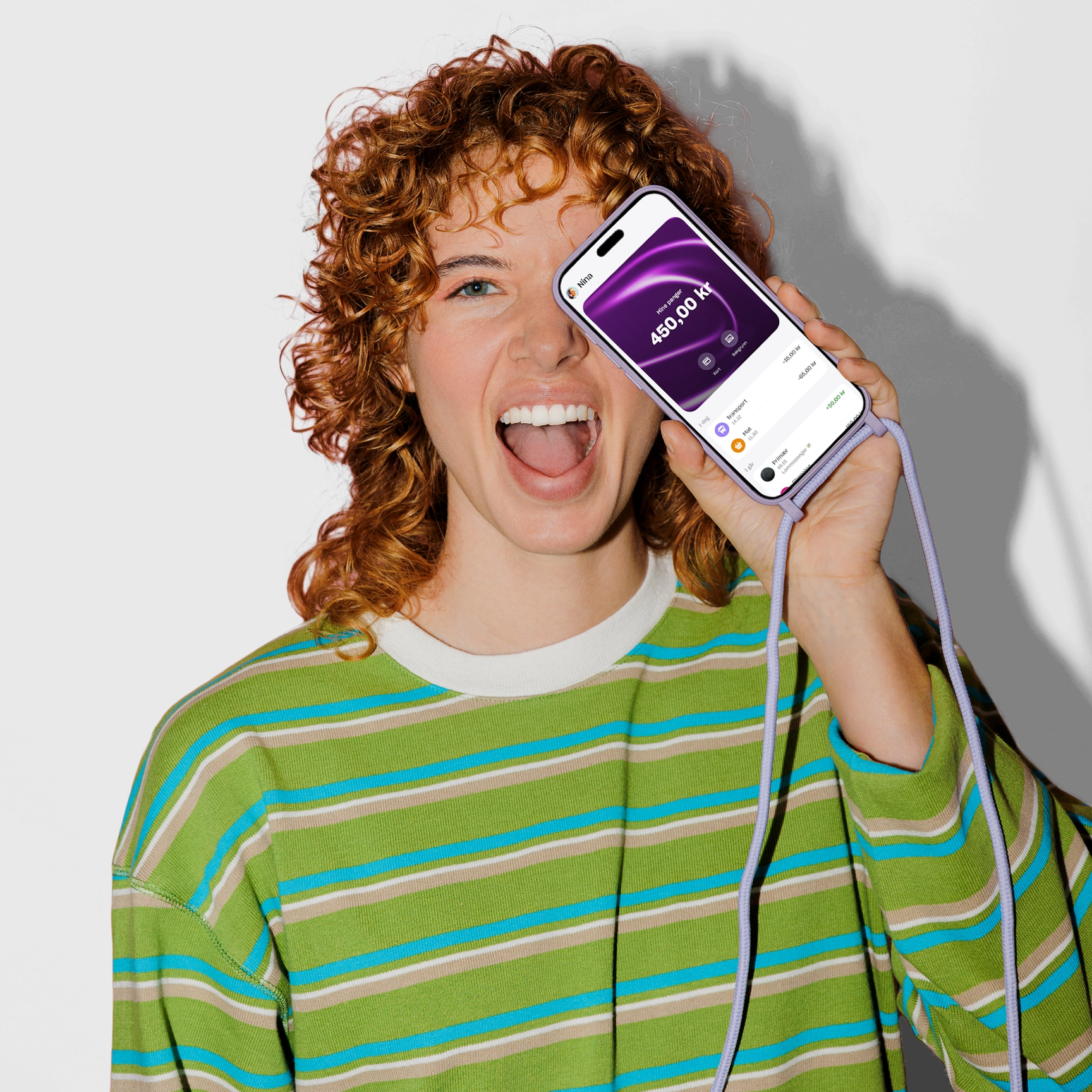 En glad ung kvinne med krøllete rødt hår holder en smarttelefon med en lilla snor opp til ansiktet, som viser en saldo på 450,00 kr på skjermen.