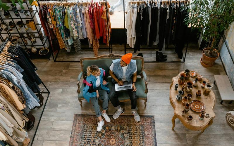 Two people sit on a couch in a vintage clothing store in Gatlinburg, one using a laptop. Racks of colorful clothes and a table with ceramics nearby create a cozy vibe—perfect for unique Gatlinburg shopping adventures. The scene is viewed from above.
