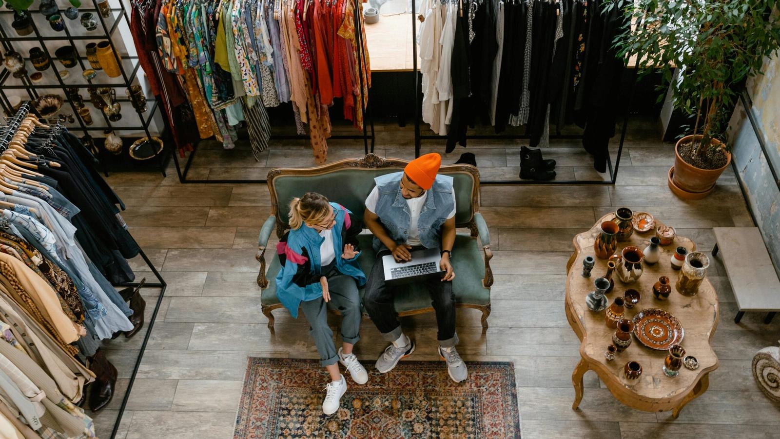 Two people sit on a couch in a vintage clothing store in Gatlinburg, one using a laptop. Racks of colorful clothes and a table with ceramics nearby create a cozy vibe—perfect for unique Gatlinburg shopping adventures. The scene is viewed from above.