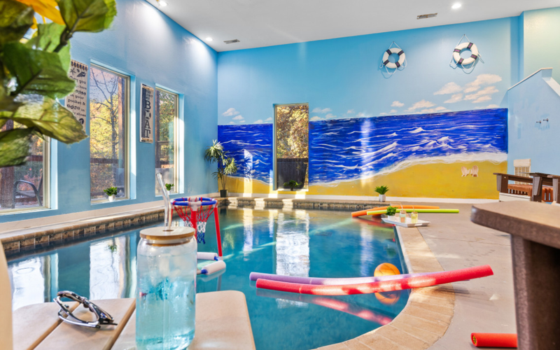 Indoor pool with a beach mural, pool toys, and seating area.