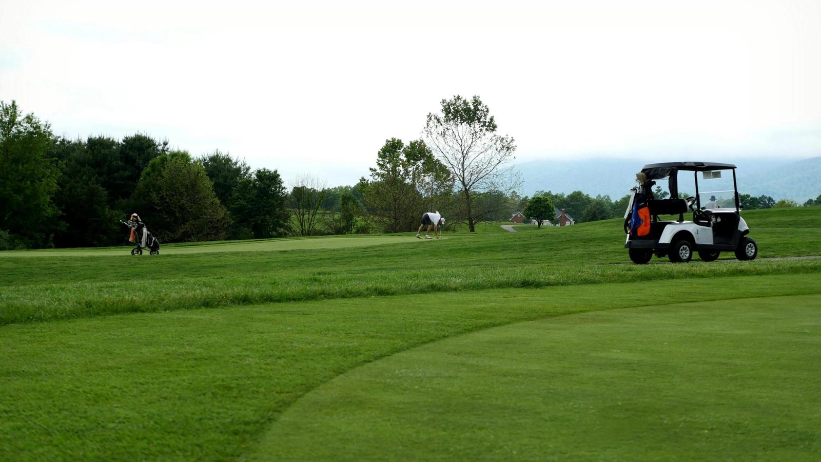 A golf course with several golfers playing in the distance. A white golf cart with clubs is parked on the grass near the right side of the image at Gatlinburg Golf Course, bordered by trees under a cloudy sky.