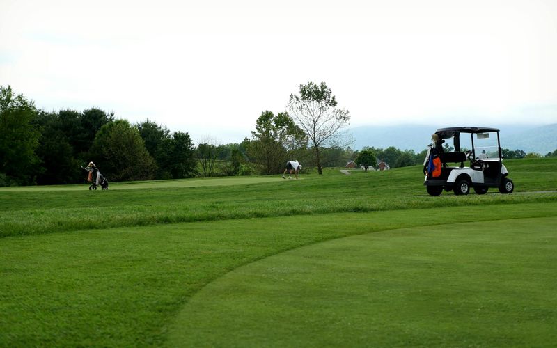 A golf course with several golfers playing in the distance. A white golf cart with clubs is parked on the grass near the right side of the image at Gatlinburg Golf Course, bordered by trees under a cloudy sky.