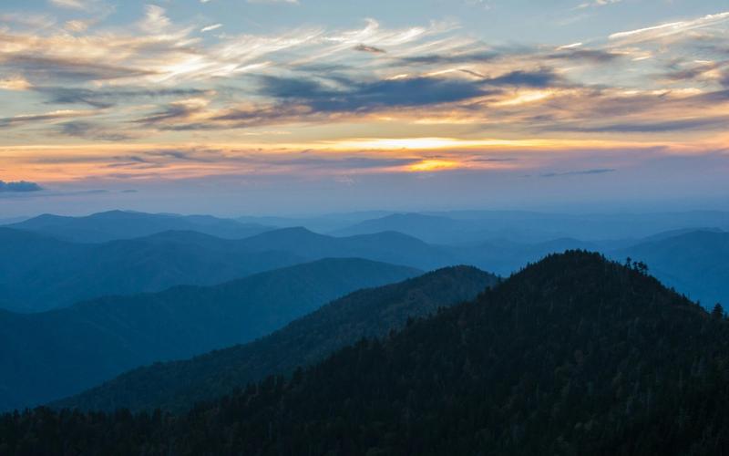 Sunset over a range of blue-tinted mountains near Gatlinburg, with layered ridges and a sky filled with scattered clouds and soft orange and yellow hues—a breathtaking scene among the top things to do in Gatlinburg.