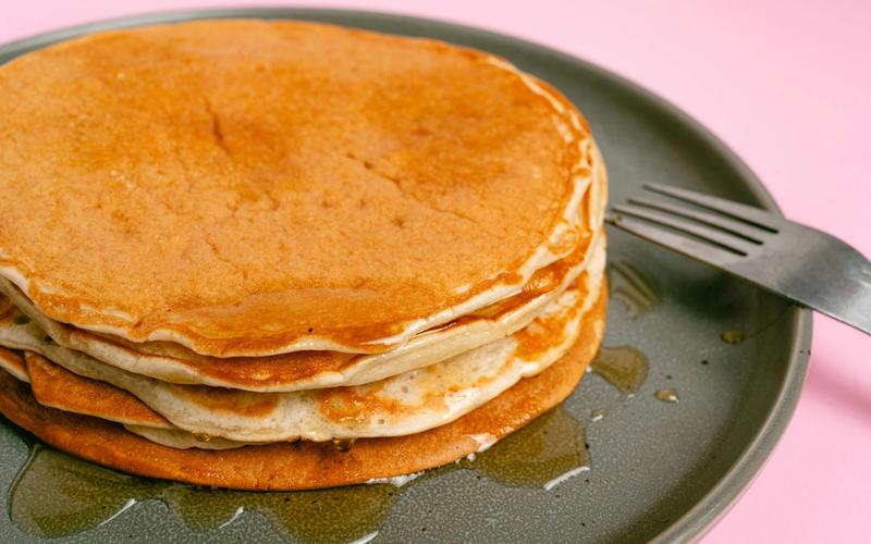 A stack of pancakes with syrup sits on a gray plate next to a fork, against a pink background, inspired by the menu at Crockett’s Breakfast Camp.