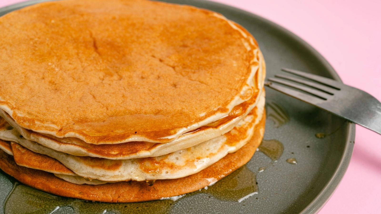 A stack of pancakes with syrup sits on a gray plate next to a fork, against a pink background, inspired by the menu at Crockett’s Breakfast Camp.