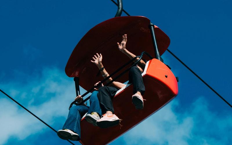Two people sit in a bright red chairlift at Gatlinburg SkyPark against a blue sky, waving and dangling their legs as they enjoy the ride—a perfect moment when visiting Gatlinburg.