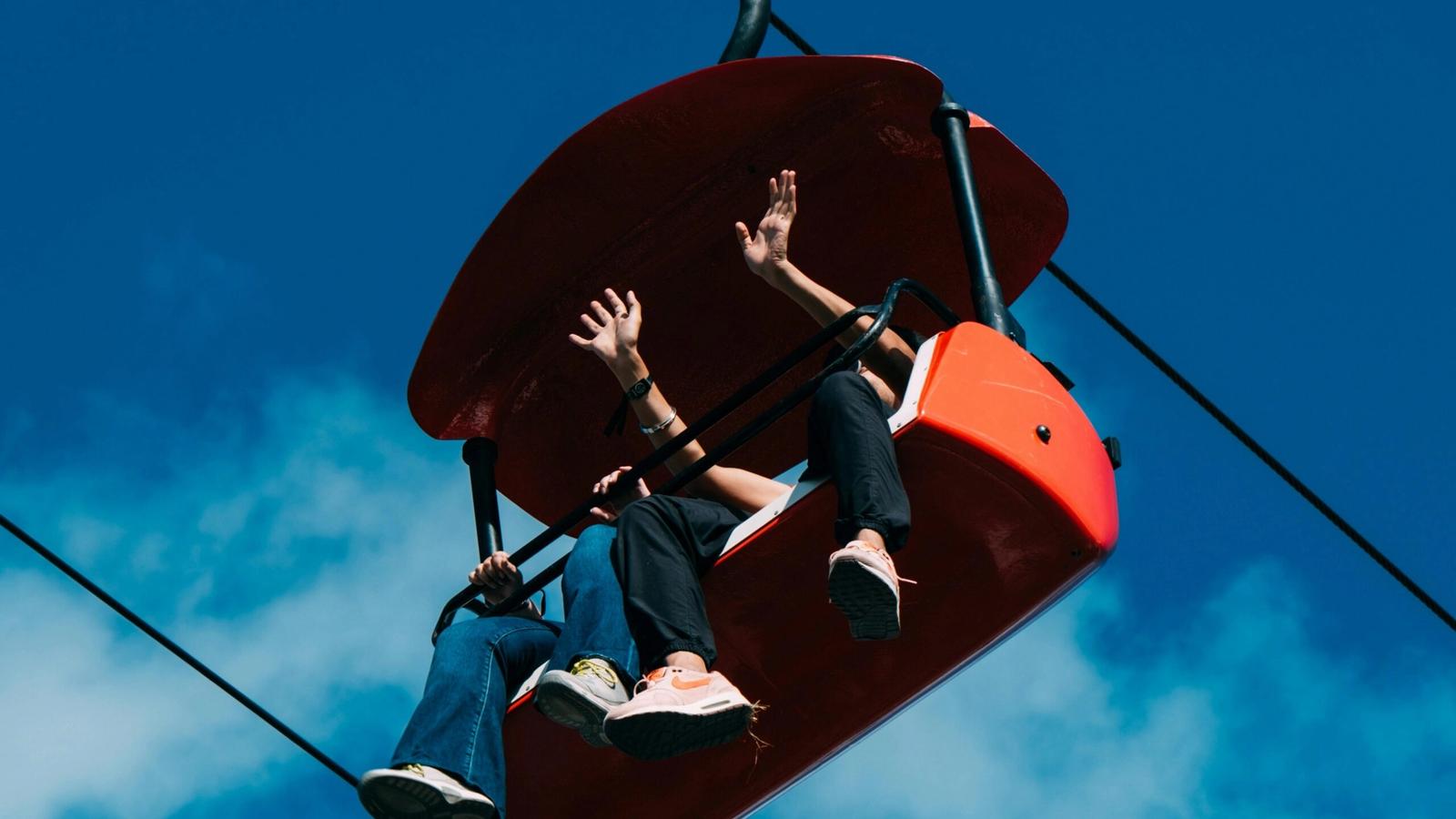 Two people sit in a bright red chairlift at Gatlinburg SkyPark against a blue sky, waving and dangling their legs as they enjoy the ride—a perfect moment when visiting Gatlinburg.