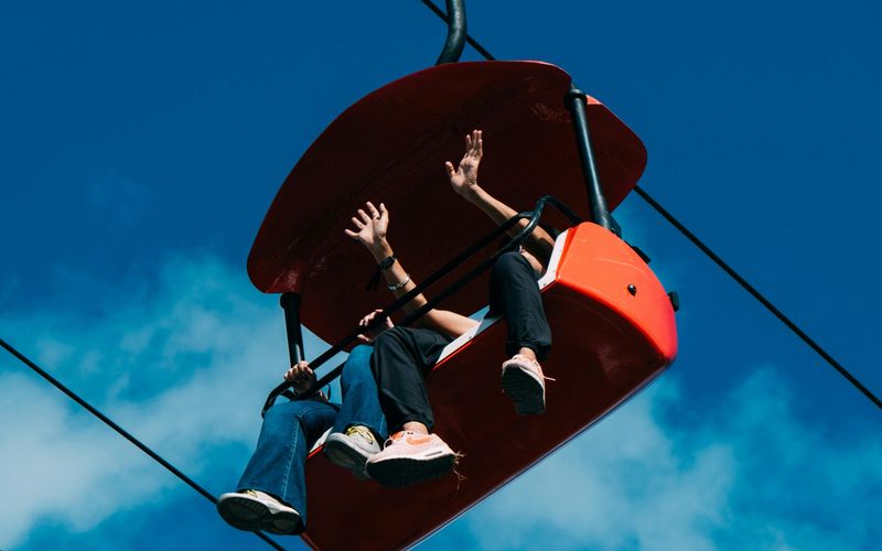Two people sit in a bright red chairlift at Gatlinburg SkyPark against a blue sky, waving and dangling their legs as they enjoy the ride—a perfect moment when visiting Gatlinburg.