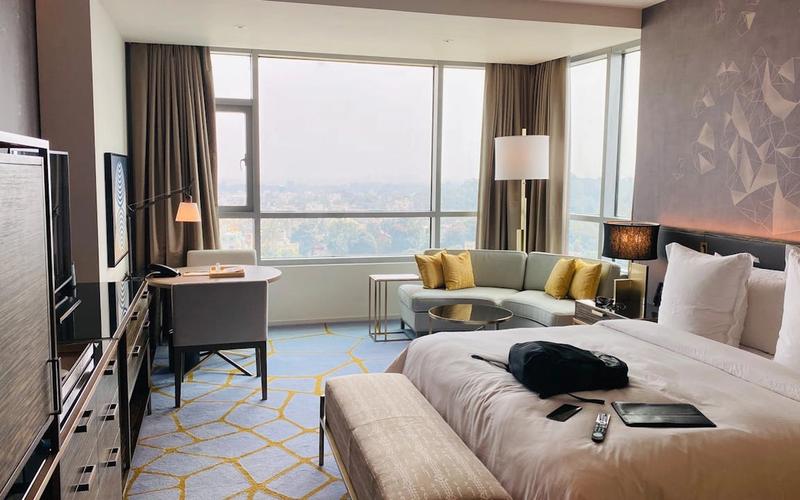A modern luxury hotel room in Downtown Gatlinburg features a large bed, light-colored bench, sofa with yellow pillows, desk by the window, and cityscape views of the Strip. A TV, remote, and black bag are also visible in the room.