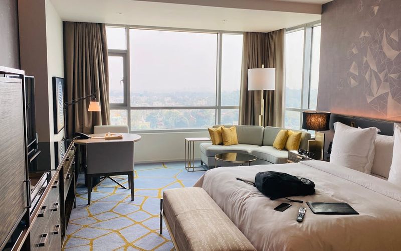 A modern luxury hotel room in Downtown Gatlinburg features a large bed, light-colored bench, sofa with yellow pillows, desk by the window, and cityscape views of the Strip. A TV, remote, and black bag are also visible in the room.