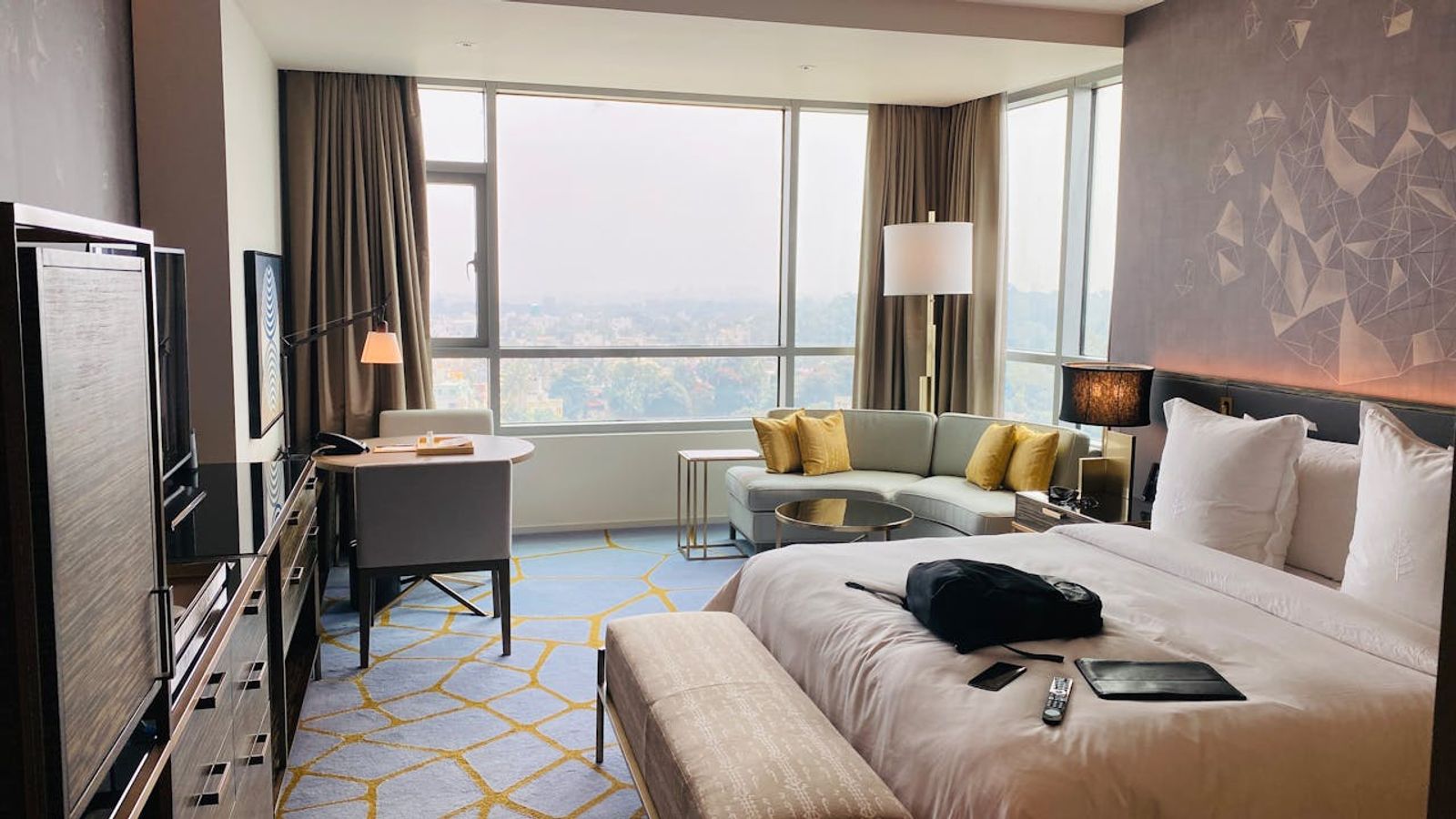 A modern luxury hotel room in Downtown Gatlinburg features a large bed, light-colored bench, sofa with yellow pillows, desk by the window, and cityscape views of the Strip. A TV, remote, and black bag are also visible in the room.