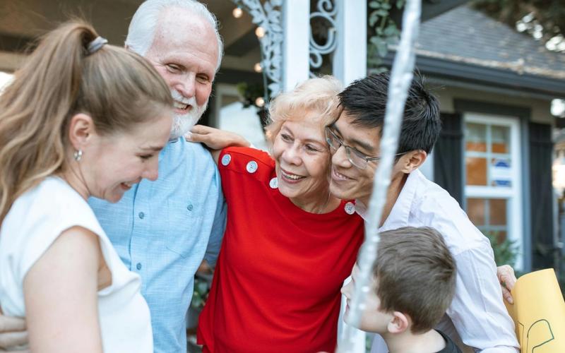 A group of five people, including two older adults, two younger adults, and a child, share a close, joyful hug outdoors in front of a house—smiling and enjoying each other’s company after playing Fun for Everyone family reunion games.
