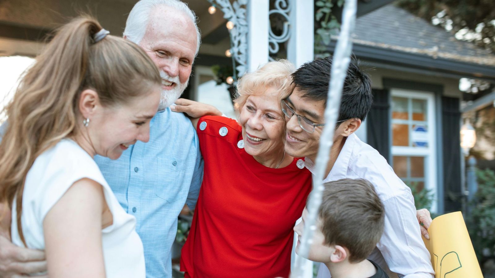 A group of five people, including two older adults, two younger adults, and a child, share a close, joyful hug outdoors in front of a house—smiling and enjoying each other’s company after playing Fun for Everyone family reunion games.