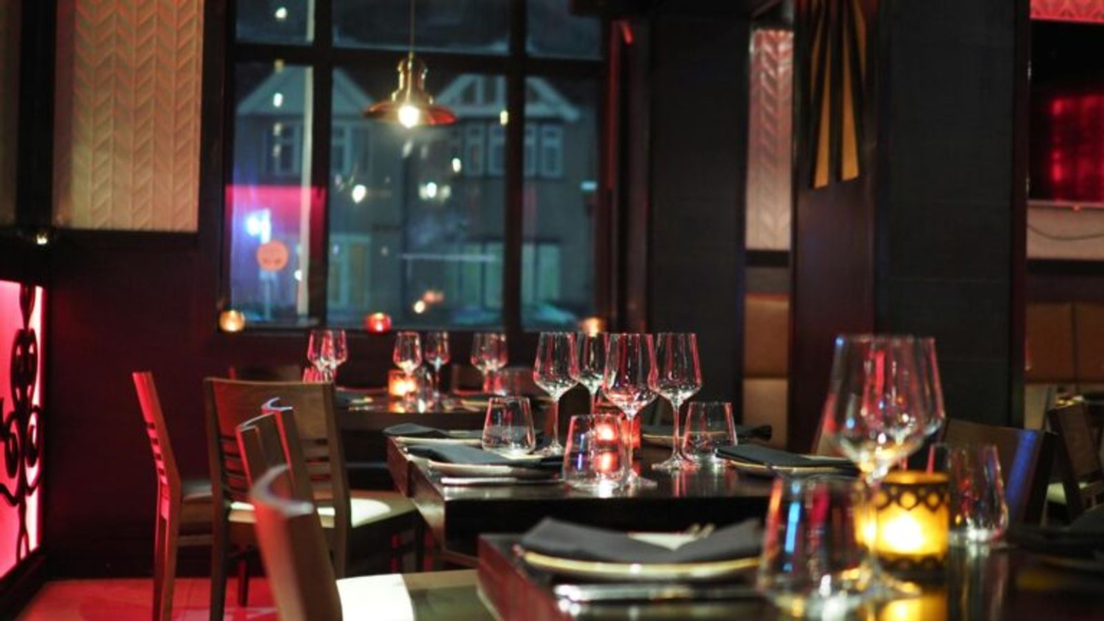 A dimly lit restaurant with tables set with wine glasses, napkins, and candles. The cozy, inviting atmosphere makes it one of the best restaurants Gatlinburg has to offer, with large windows framing views of buildings outside.