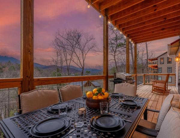 A covered wooden deck with a dining table set for four, featuring black plates and a bowl of citrus fruit. Cozy seats line the table, and string lights hang above. Bare trees and mountains are visible under a vibrant sunset sky.