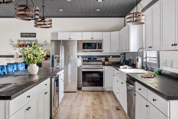 Modern kitchen with white cabinets, stainless steel appliances, black countertops, a farmhouse sink, wooden floor, and decorative light fixtures. A vase of flowers sits on the island, and a Coffee Bar sign is on the wall.