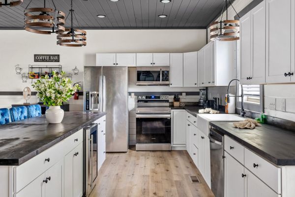 Modern kitchen with white cabinets, stainless steel appliances, black countertops, a farmhouse sink, wooden floor, and decorative light fixtures. A vase of flowers sits on the island, and a Coffee Bar sign is on the wall.