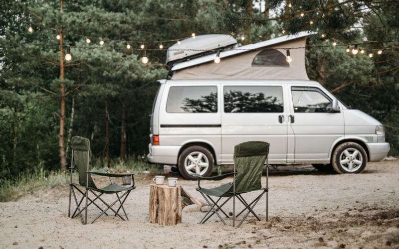 Two camping chairs and a small table with mugs sit on sandy ground in front of a campervan beneath hanging string lights, surrounded by pine trees—perfect for camping near Gatlinburg and enjoying the tranquility of the outdoors.