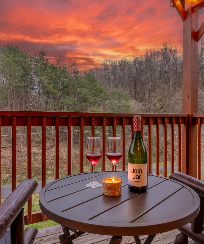 A round table on a wooden porch holds two glasses of red wine, a lit candle, and a Jam Jar wine bottle. Two chairs face a scenic sunset with vivid orange and purple skies over a forested landscape.